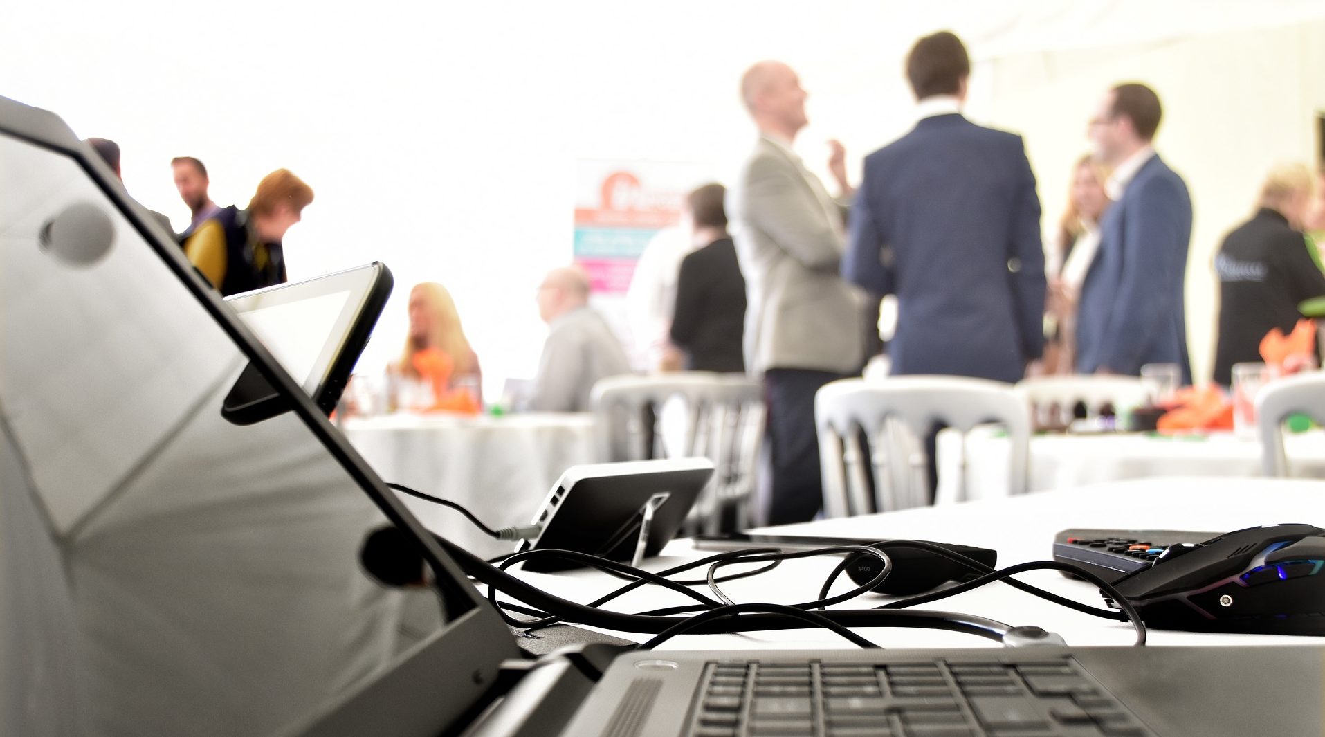 Laptop and equipment on a table with focused foreground, blurry background showing millennials networking in a white tent event space.