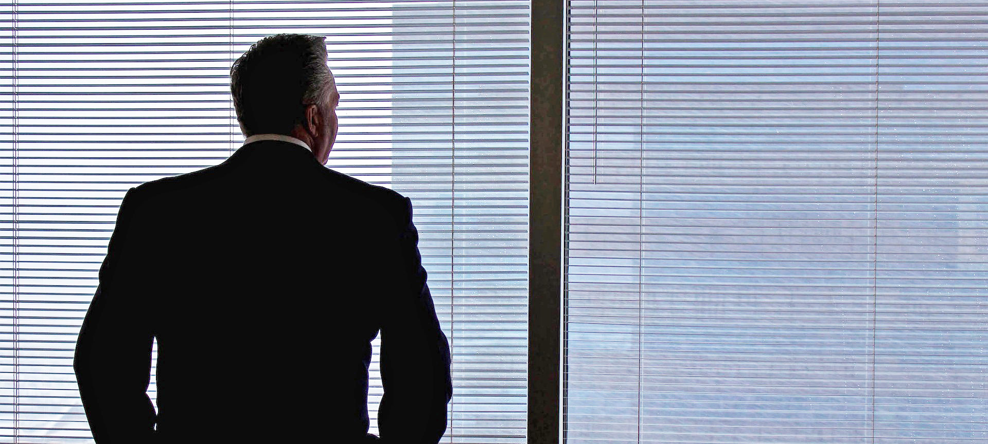 A man in a business suit stands in front of a window with closed blinds, facing away from the camera, contemplating succession planning.