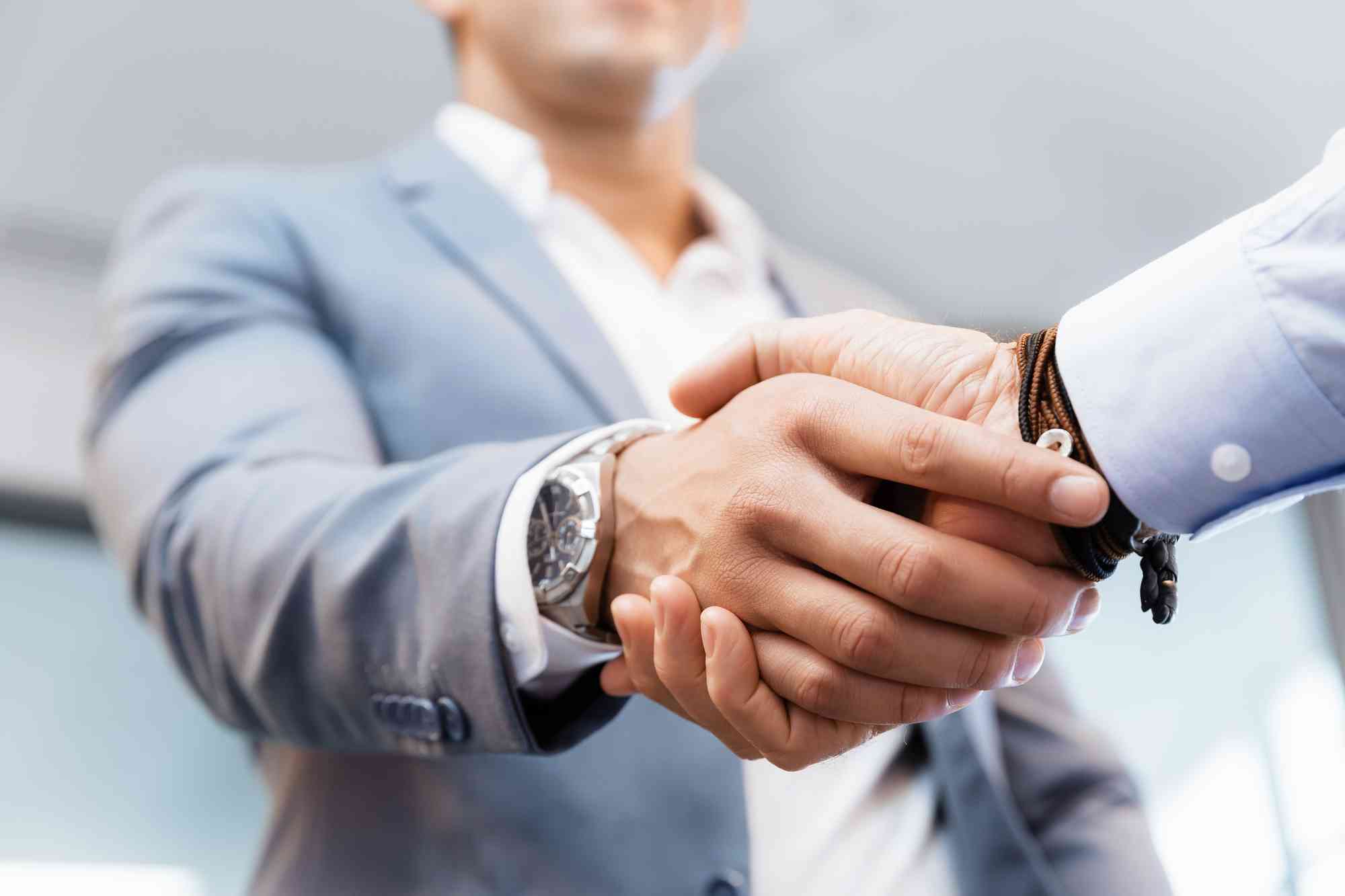 Two business professionals shaking hands, focus on hands with blurred office background, emphasizing a formal agreement or greeting in their functional specialties.