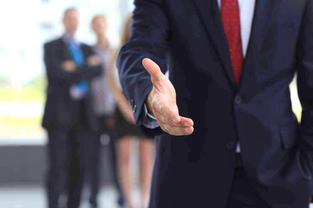 Close-up of an experienced professional extending a handshake, with blurred colleagues in the background.