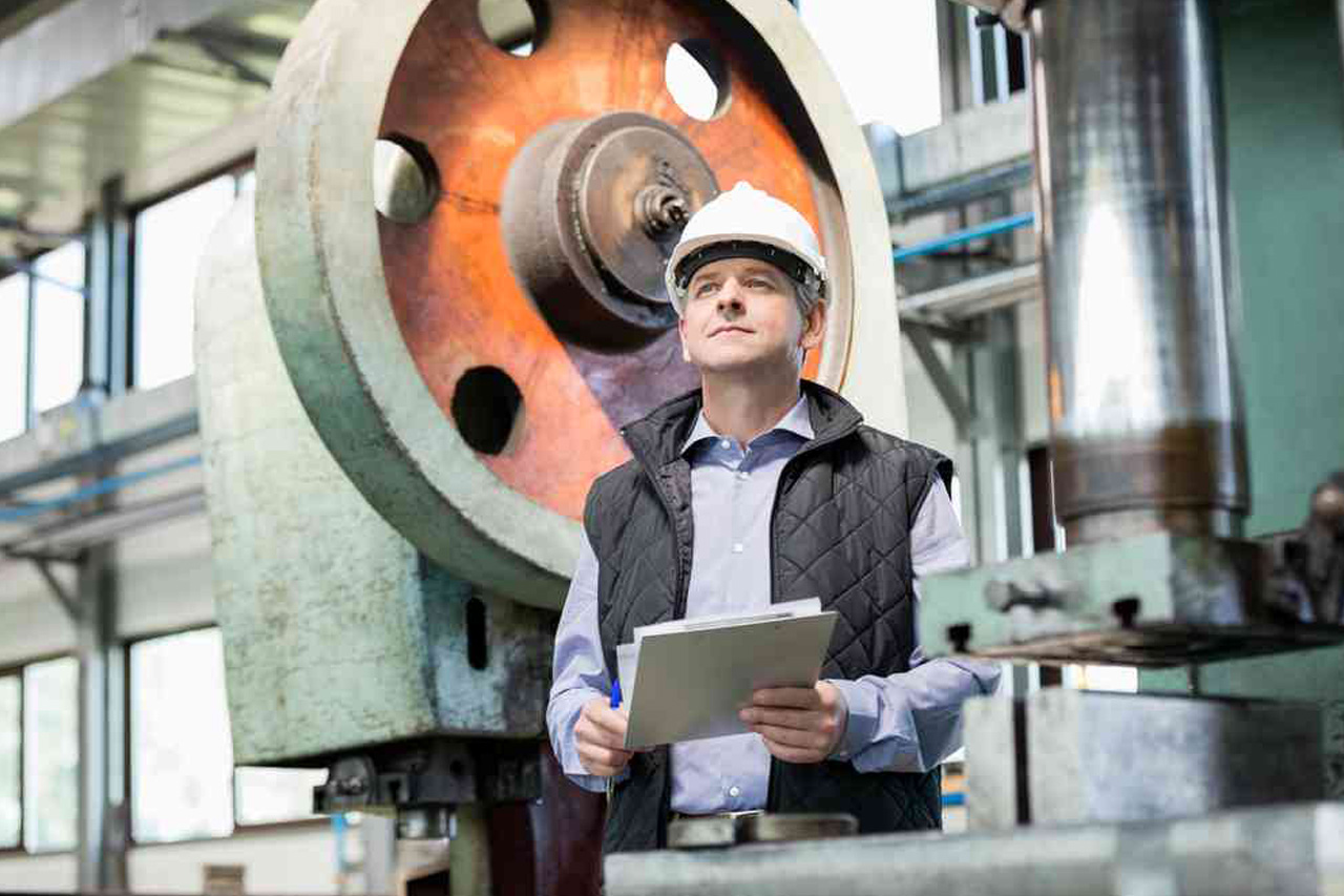Engineer in hard hat inspecting machinery in an industrial plant, holding a tablet and a clipboard for description analysis.