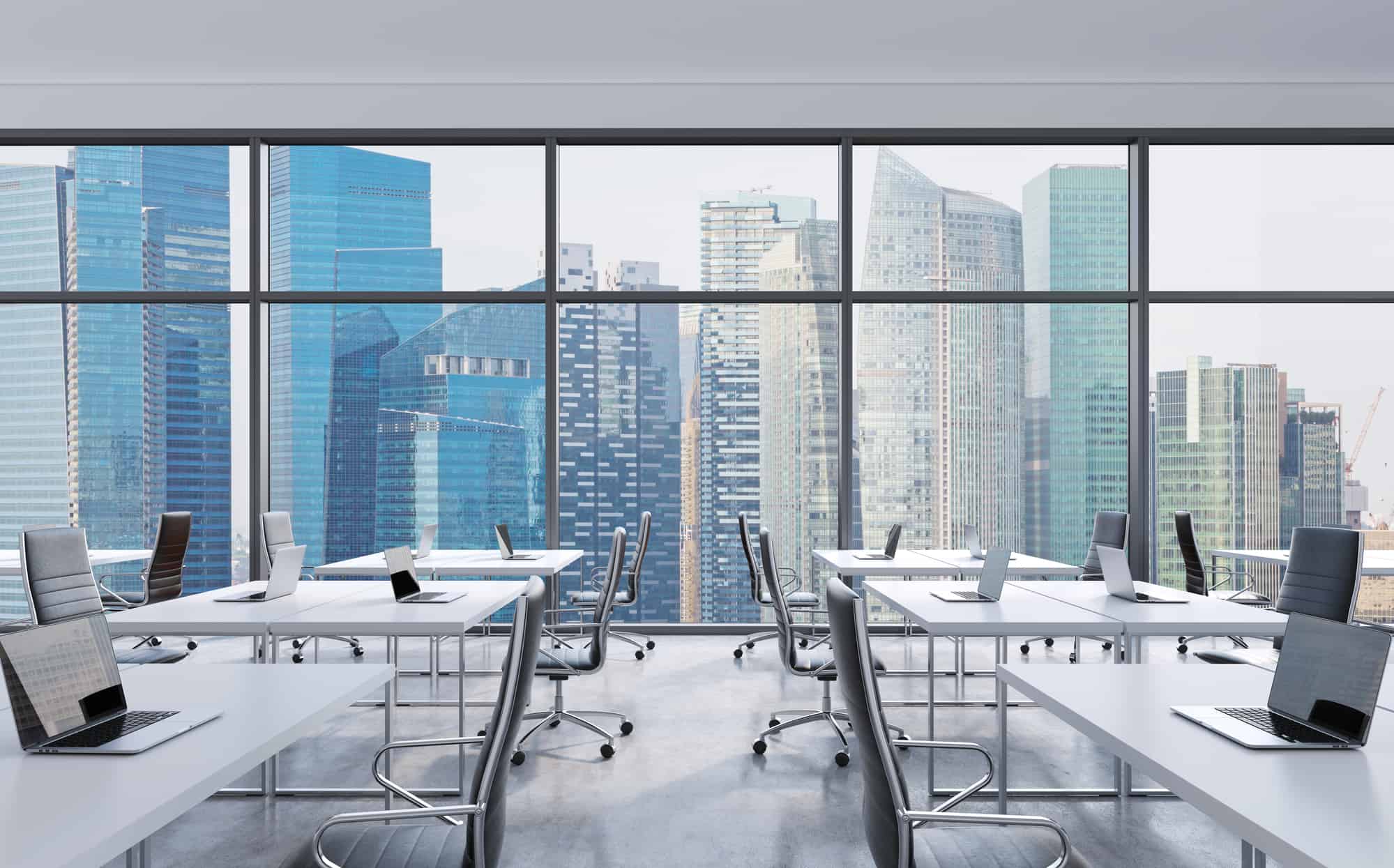 Modern office space with rows of white desks and laptops, owned by CEOs, overlooking a cityscape with skyscrapers through large windows.