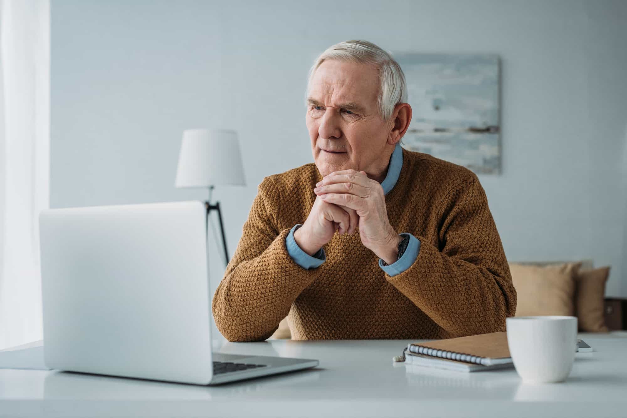 Elderly man in a sweater sitting at a desk, looking thoughtfully at his laptop in a home office setting.