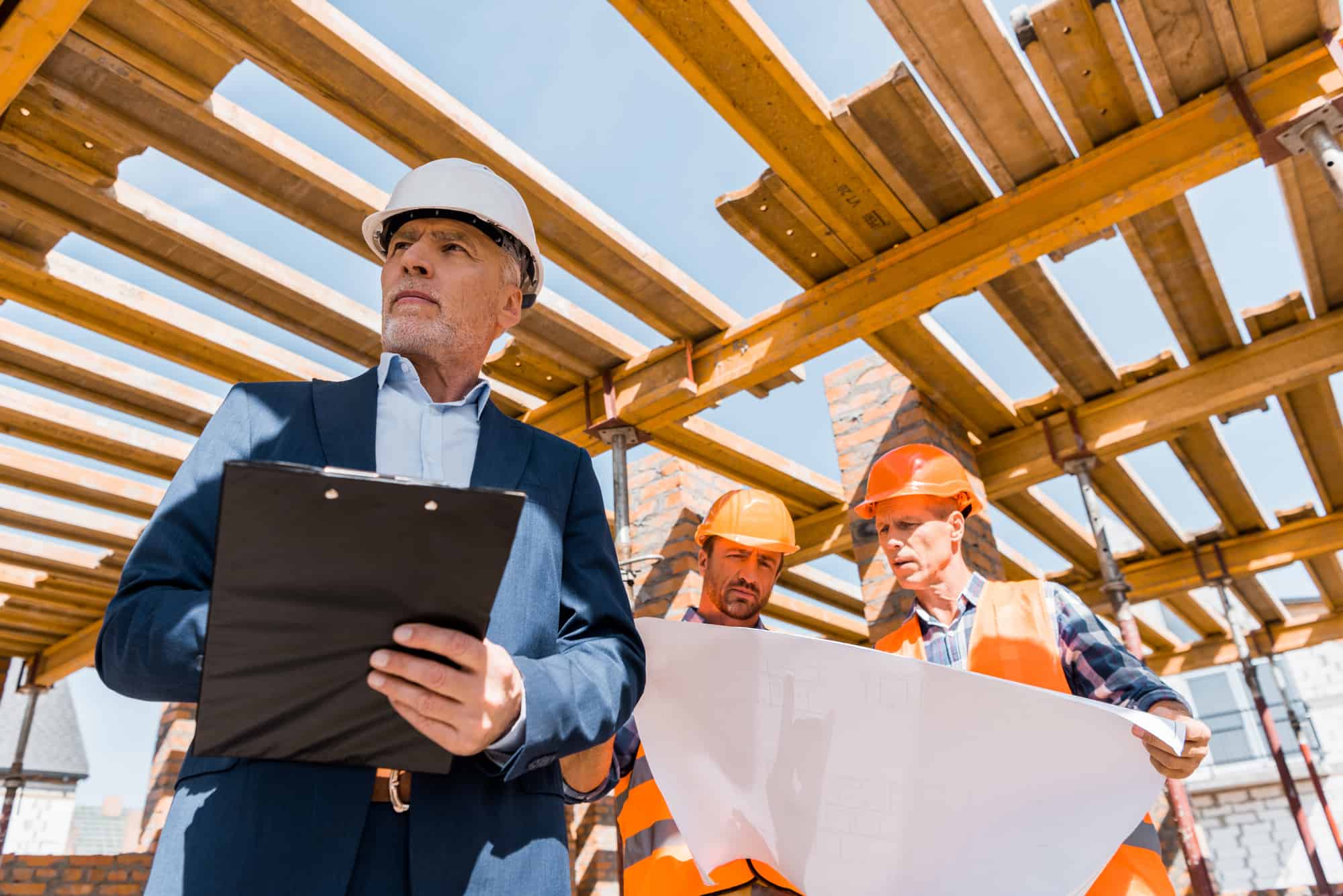 Three construction workers, wearing hard hats and addressing the skilled labor shortage at a construction site, with one man in the foreground holding a clipboard.