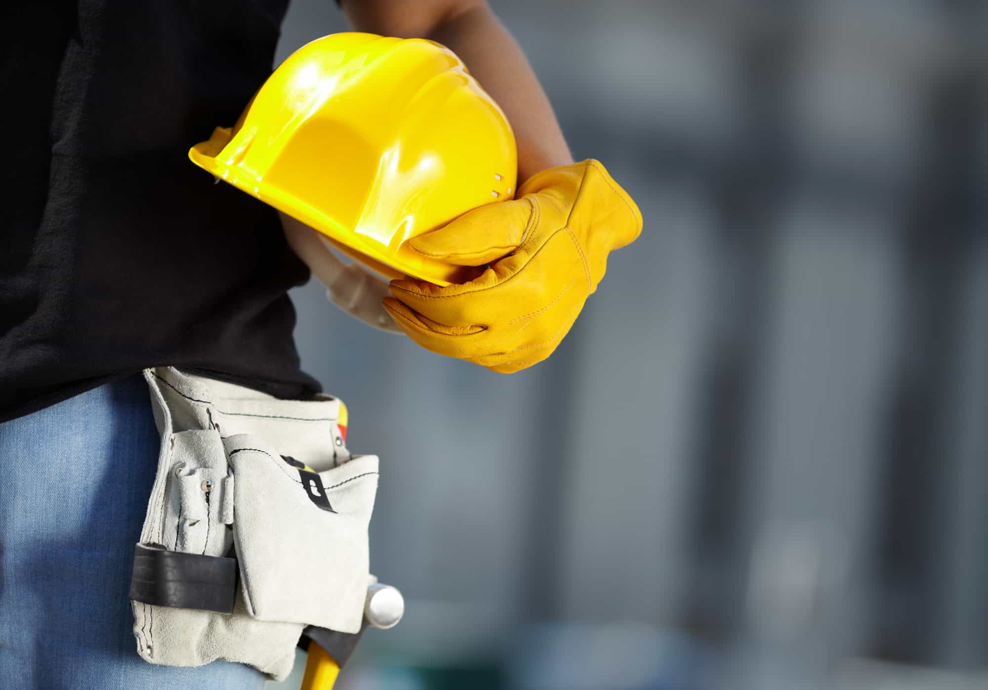 Close-up of a construction worker holding a yellow hard hat and gloves, with a tool belt around their waist, blurred background of a construction site in the construction industry.