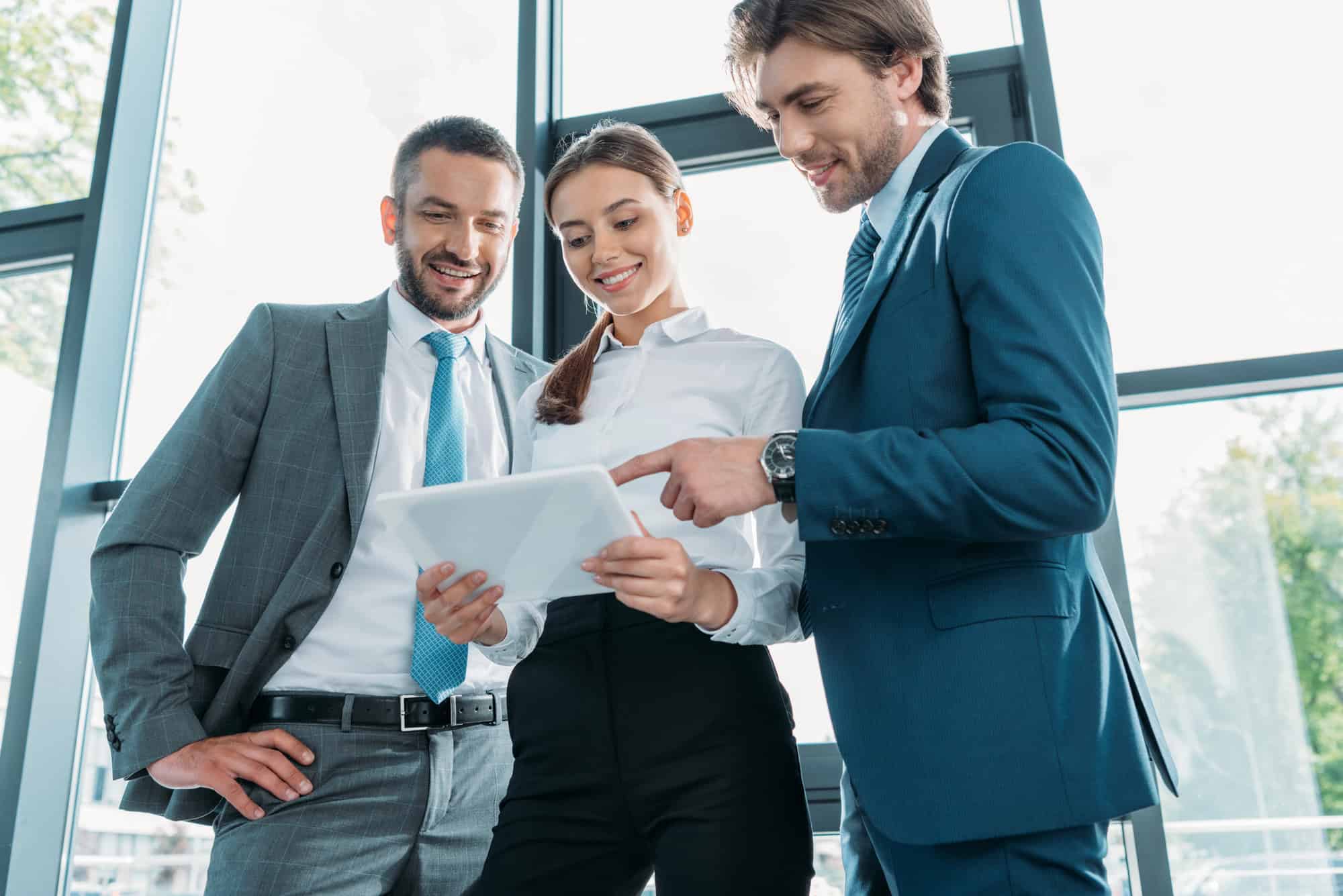 Three business professionals, two men and one woman, review a document on a tablet together. They are standing in an office with large windows and natural light, embodying the leadership characteristics that define a good leader in 2024.