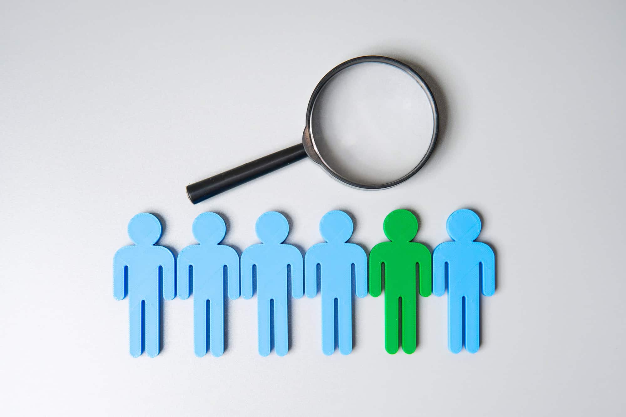 A magnifying glass is positioned over a green human figure among five blue human figures, symbolizing talent acquisition and attention to emerging trends.