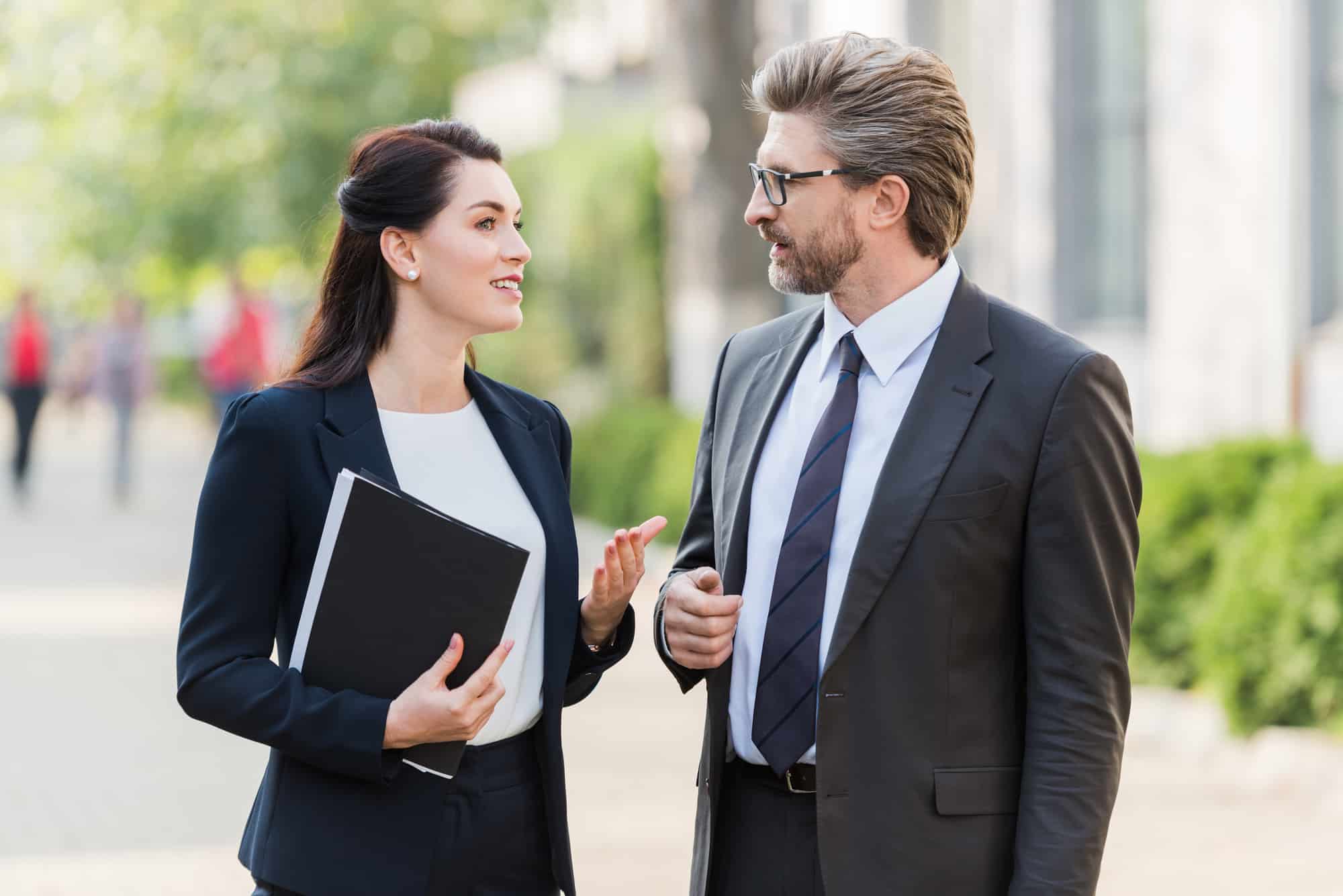 Two business professionals in formal attire are walking outdoors, engaged in a conversation about a CEO executive search. The woman holds a folder as trees and people can be seen in the background.