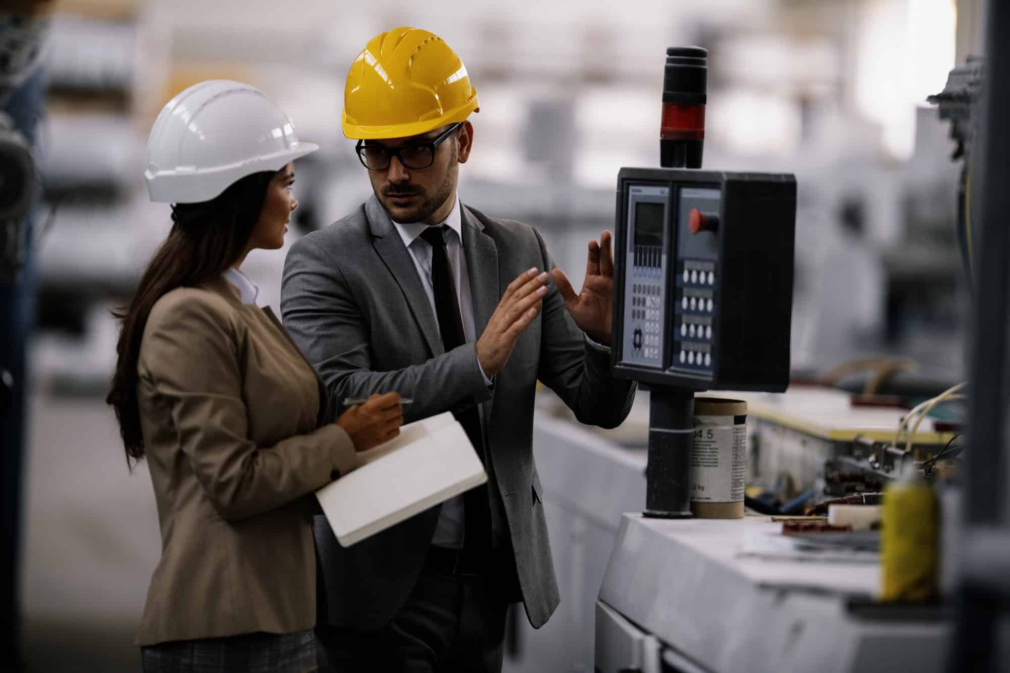 Two engineers in hard hats engage in discussion while examining an industrial control panel, one jotting notes. Their detailed analysis mirrors the precision seen in a CIO executive search.