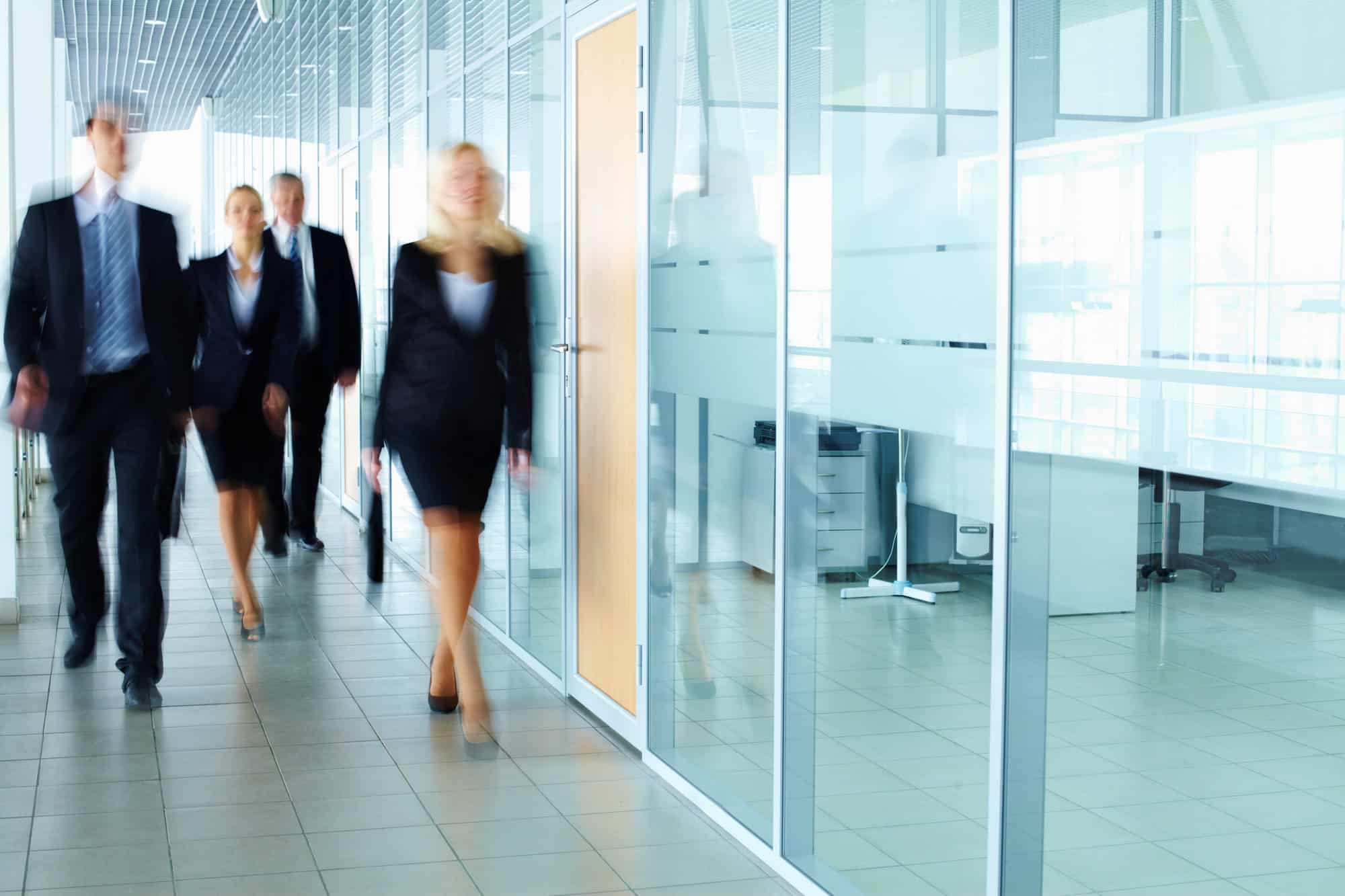 Office hallway with people in business attire walking past glass-walled rooms, where the Chief People Officer engages with colleagues, highlighting organizational differences to the attentive team.