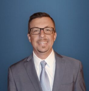 A man from our team, wearing glasses, a suit, white shirt, and tie, smiles confidently in front of a plain blue background.