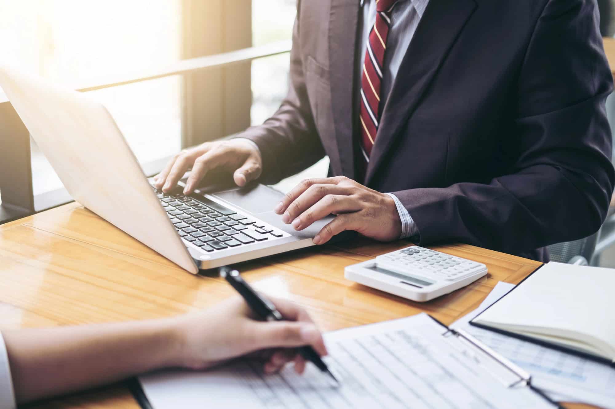 Two people in business attire work at a desk with a laptop, calculator, and documents. One types on the laptop while the other writes notes on paper, analyzing 2025 executive compensation trends and C-Suite pay structures.