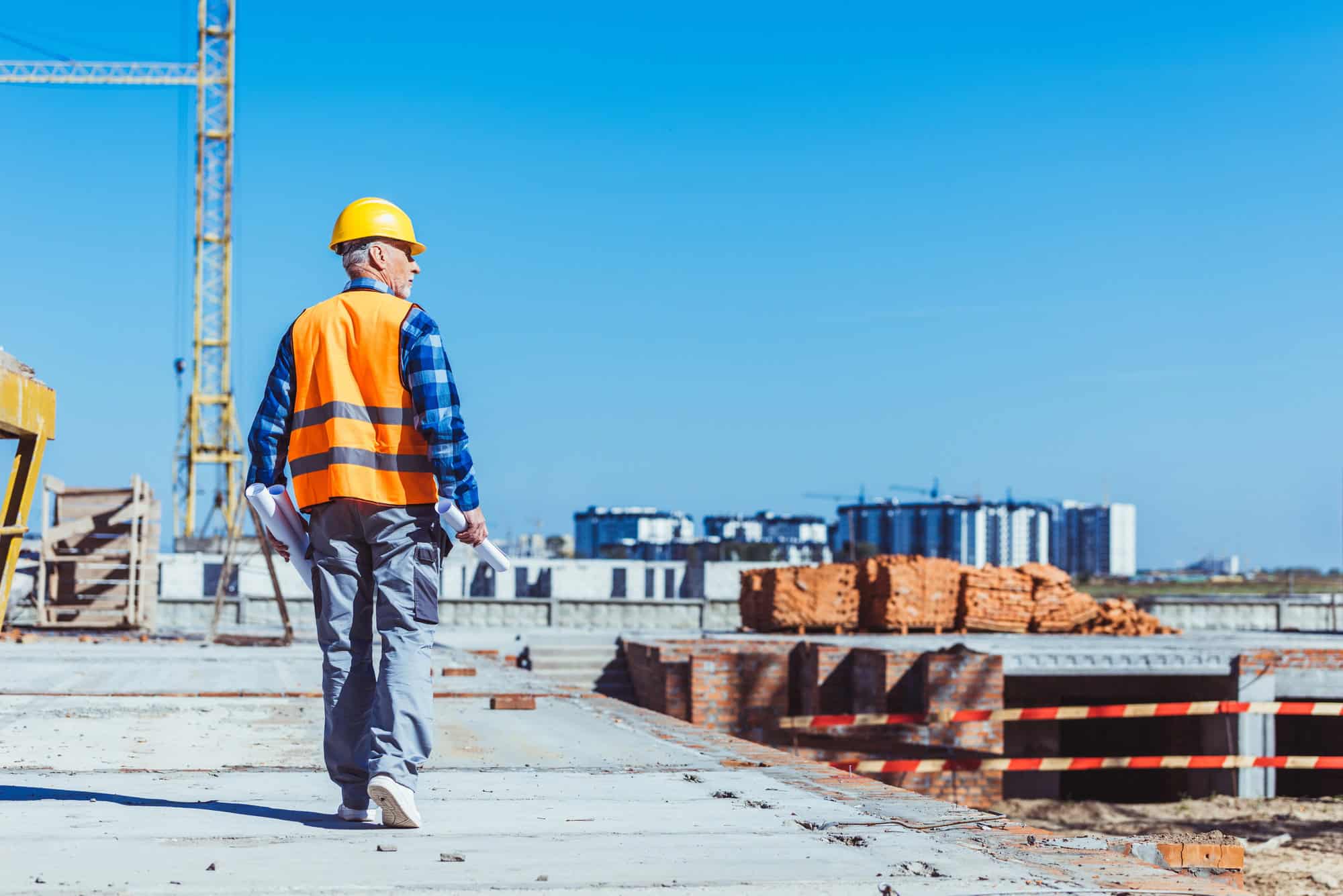 A construction worker in a hard hat and safety vest walks on a building site, embodying construction leadership as he holds rolled-up plans, with cranes and materials visible in the background.