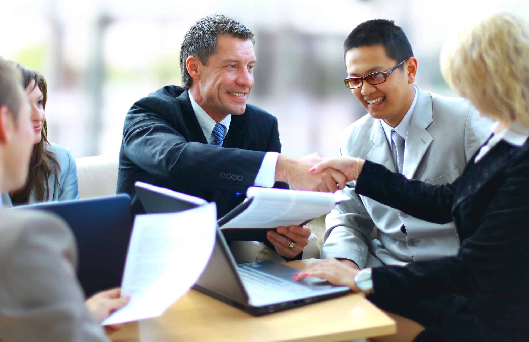 Four business professionals in suits are meeting at a table with documents and a laptop; two of them are shaking hands and smiling.