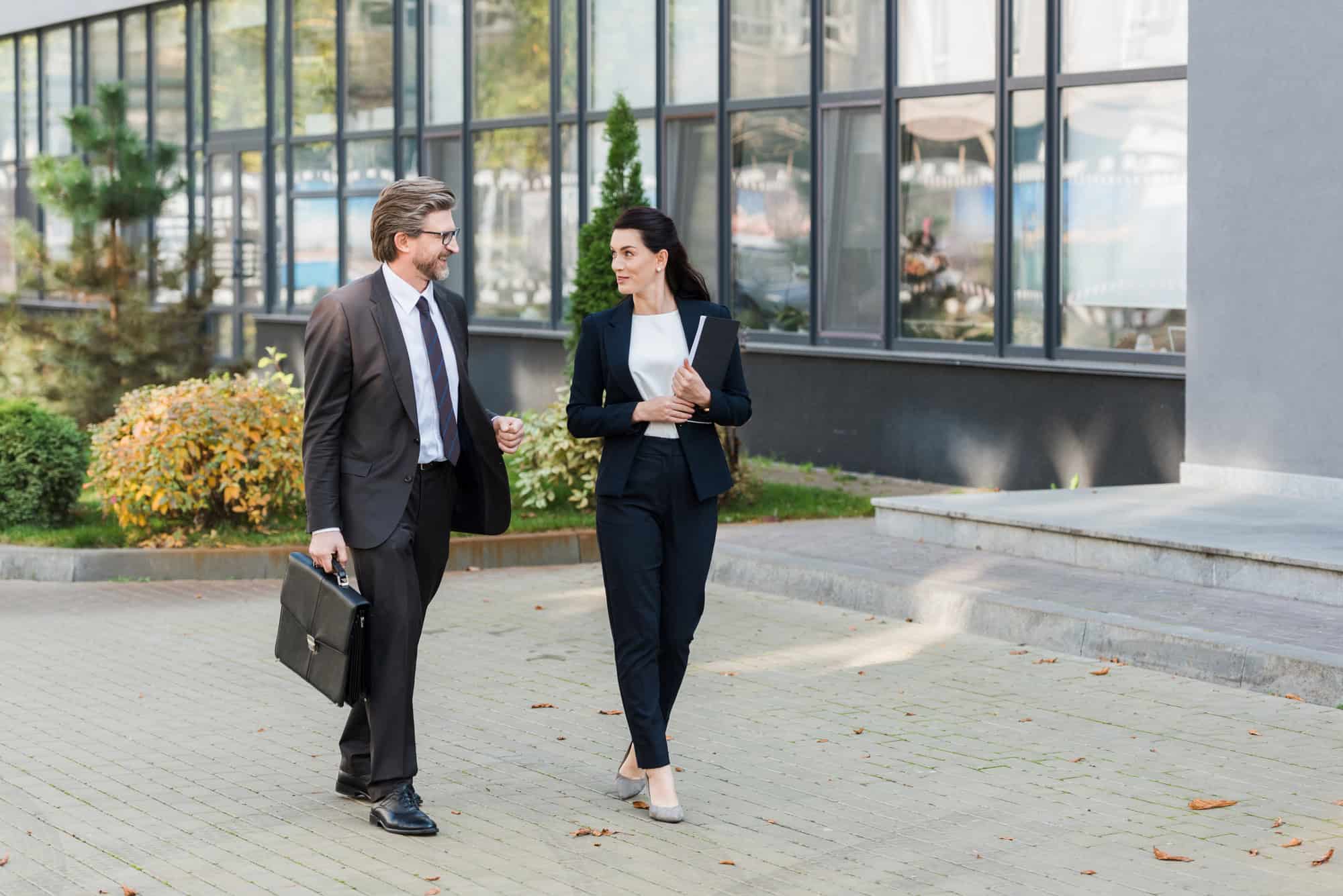 Two business professionals in suits walk and talk outside an office building; one carries a briefcase, the other holds a folder—perhaps discussing silent risk or leadership gaps facing their CFO.