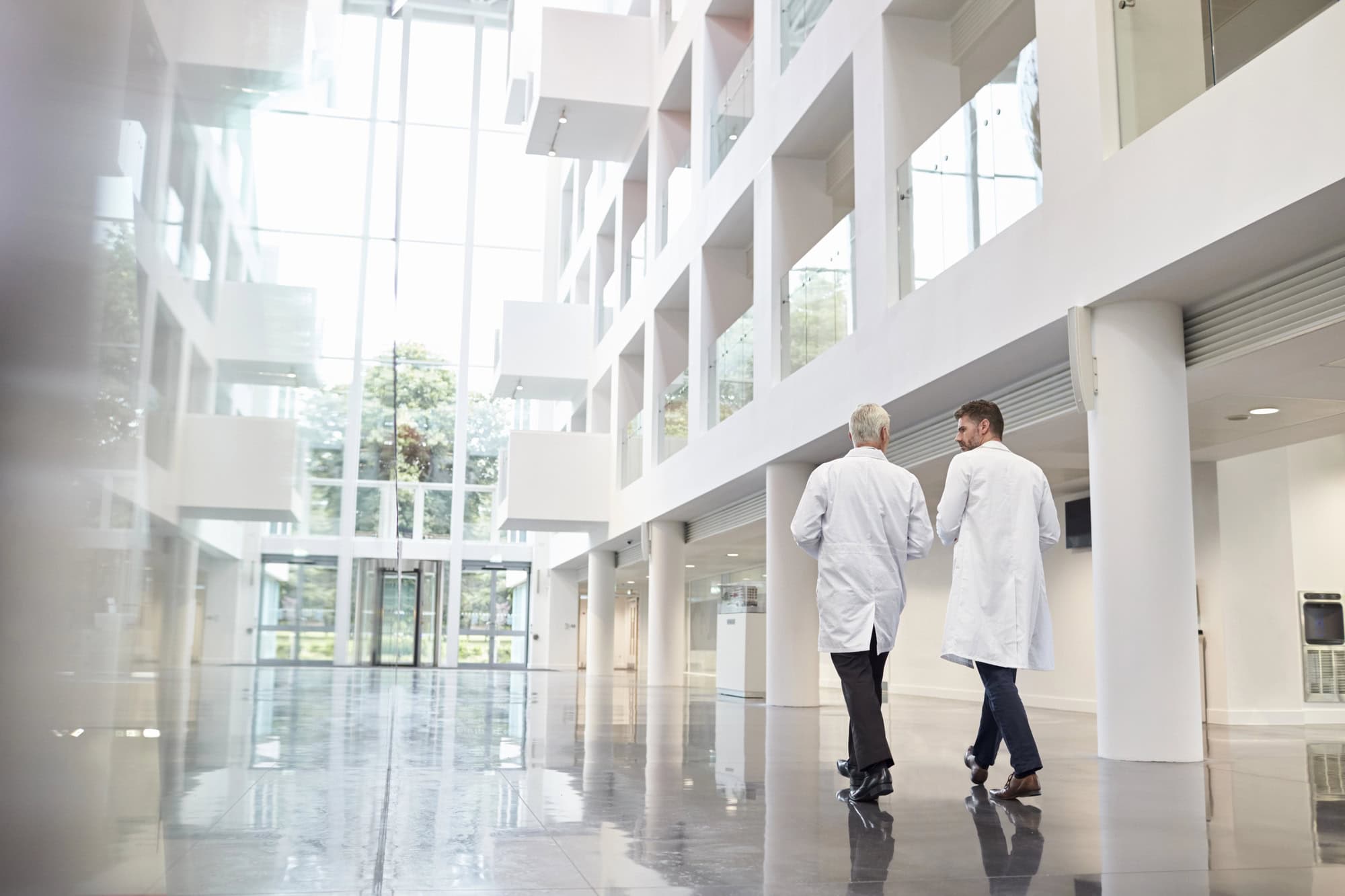 Two people in white lab coats walk through a modern, bright building with large windows and a shiny floor—an inspiring setting for Pharma Leadership 2025 and C-Suite Pharma Talent to shape the future of healthcare innovation.