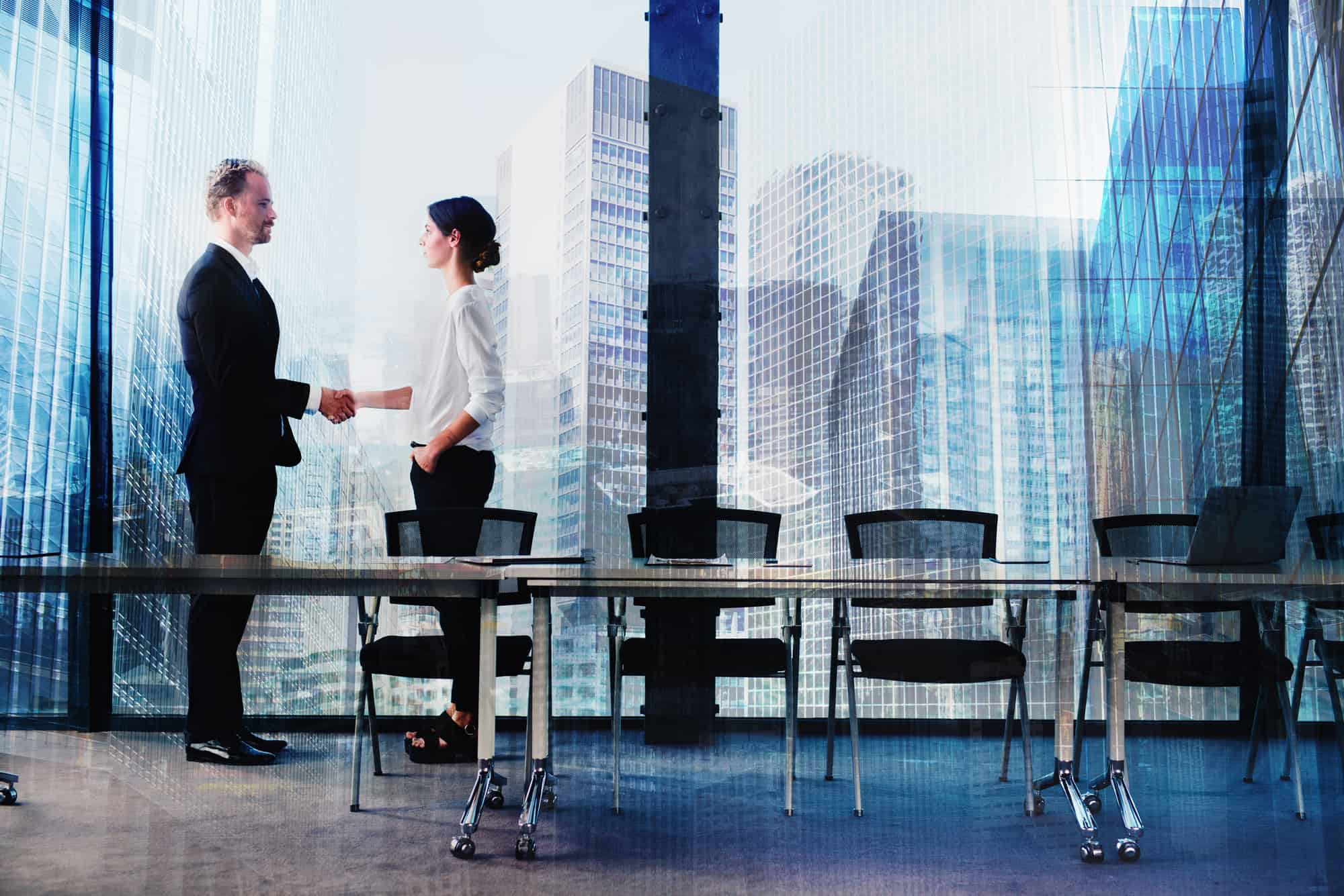 Two business professionals shake hands in a modern conference room with large windows overlooking a city skyline.